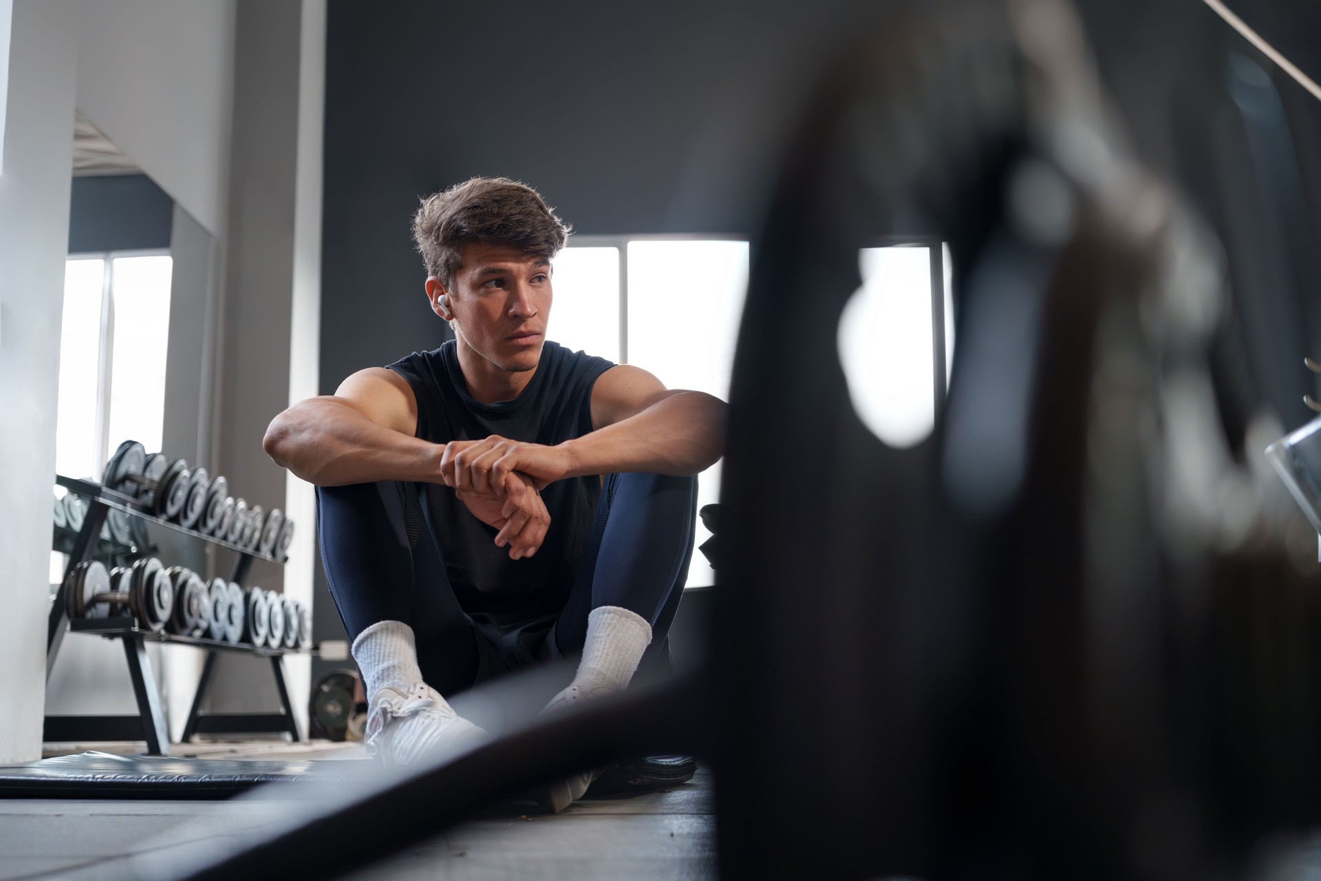 Junger athletischer Mann, der auf dem Boden des Fitnessstudios sitzt, sich ausruht und nach dem Training nachdenklich aussieht